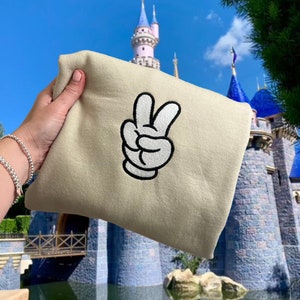 K&ouml;nnte beinhalten: Ein beiger Pullover mit einer wei&szlig;en gestickten Mickey Mouse Hand, die das Friedenszeichen macht.
