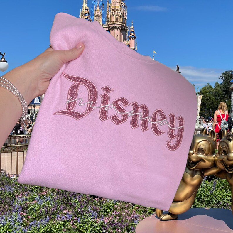 K&ouml;nnte beinhalten: Rosafarbener Pullover mit einem glitzernden pinkfarbenen "Disney"-Motiv in gotischer Schrift. Der Pullover wird vor einem Schloss gehalten.