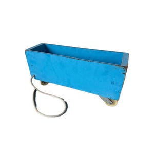May include: A rectangular, light blue wooden cart with four small wheels and a white rope handle. The cart is empty and appears to be vintage or antique, with some signs of wear and tear.