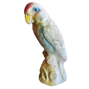 May include: A vintage parrot figurine, perched on a tree stump. The bird has a red head, blue eyes, and a yellow beak. The body is primarily off-white with hints of blue and green. The base is a muted yellow.