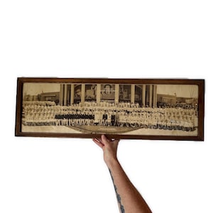 May include: A framed black and white photograph of a large group of people, likely a school or organization, posed in front of a building with columns. The photograph is in a dark wooden frame.