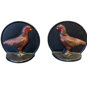 May include: Two cast iron rooster doorstops. Each doorstop features a brown rooster with a red comb and yellow beak, set against a black circular background. The base is a dark gray color.