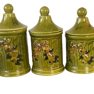 May include: Three olive green ceramic canisters with lids. Each canister features a decorative relief of fruit and leaves in shades of brown and tan. The canisters have a ribbed design and a rounded knob on top.
