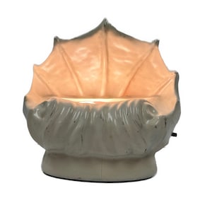 May include: A cream-colored, decorative lamp shaped like a stylized seashell. The lamp has a textured surface and a warm, glowing interior. The base is round and the lamp is plugged in.