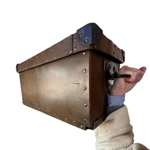 May include: A vintage brown trunk with brass rivets and dark blue corner protectors. The trunk has a black handle and is held by a person wearing a cream-colored jacket. The trunk appears to be made of wood and is rectangular in shape.