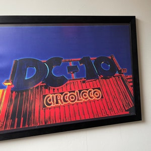Puede incluir: Un póster enmarcado con una imagen roja, amarilla y azul de un edificio con el texto "DC-10" y "CIRCOLOCO" en luces de neón.