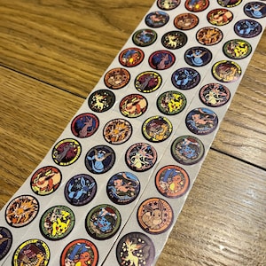 May include: A long strip of circular stickers featuring various colourful Pokémon characters. Each sticker has a distinct border and displays a different creature, including Pikachu, Eevee, Charmander, and others, with Japanese text printed on each individual design.