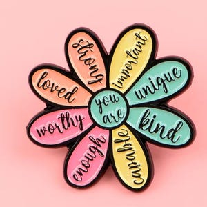 May include: A flower-shaped enamel pin with black metal outlines. The centre reads "you are" in black script. The eight petals are coloured in shades of orange, yellow, pink, and teal, each featuring the words "strong," "important," "unique," "kind," "capable," "enough," "worthy," and "loved."