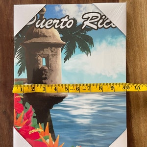 May include: A colourful canvas print featuring a vintage illustration of a stone tower with a palm tree in the background. The text "Puerto Rico" is written in a stylised font across the top of the image. The tower is situated on a cliff overlooking a blue body of water. Red flowers are in the foreground.