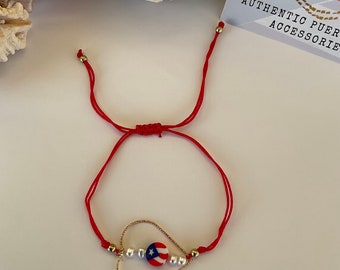 May include: Red string bracelet with a gold heart charm featuring a Puerto Rican flag charm and white pearl beads. The bracelet is tied with a knot and has gold beads on each end.