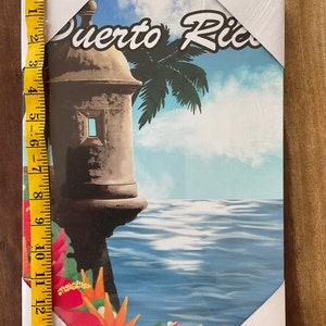 May include: A colourful canvas print featuring a view of a fort in Puerto Rico. The fort is brown and has a white window. The background shows a blue ocean, a palm tree, and a cloudy sky. The text "Puerto Rico" is written in a white, stylised font.