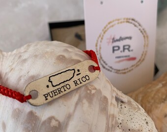 May include: Red string bracelet with a silver charm shaped like the island of Puerto Rico. The charm says "PUERTO RICO" and has a small outline of the island.