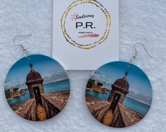 May include: A pair of round wooden earrings with a photo of a stone building with a blue roof and a view of the ocean. The earrings are silver and have a hook closure. The earrings have the text "#tradiciones P.R. PUERTO RICO" on them.