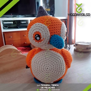 May include: A crocheted stuffed toy bird with orange, white, and blue accents. The bird has a large, round head with a small beak and large, black eyes. The bird is sitting on a wooden surface.