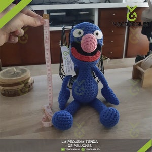 May include: A blue crocheted plush toy with a pink nose and red mouth. The toy is sitting on a wooden table with a measuring tape next to it. The toy is about 20 inches tall.