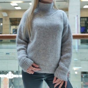 Puede incluir: Un jersey de cuello alto gris claro de gran tamaño. El jersey tiene un dobladillo y puños de canalé, y un cuello alto de canalé. La prenda de punto parece suave y cómoda, con un aspecto ligeramente texturizado. Las mangas son largas y el ajuste es holgado.