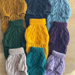 Puede incluir: Una colección de nueve suéteres tejidos para mascotas, cada uno con un patrón de trenzas y un cuello alto acanalado. Los suéteres están disponibles en varios colores sólidos, incluyendo amarillo mostaza, azul marino, azul claro, verde oscuro, morado, beige, verde azulado y lavanda.