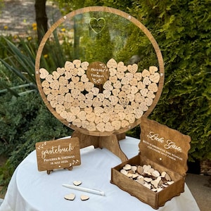 May include: A wooden wedding guest book alternative with a clear circular frame filled with heart-shaped wooden tags. The frame has a wooden base and a small sign that says "Paulina & Thomas". A wooden box and sign are also included.