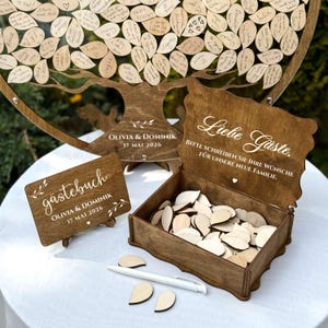 May include: A wooden guest book set for a wedding. The set includes a tree-shaped guest book, a small sign, a box with wooden leaf-shaped tags, and a pen. The tree has leaf-shaped tags with handwritten messages. The box has the text "Liebe G&auml;ste."