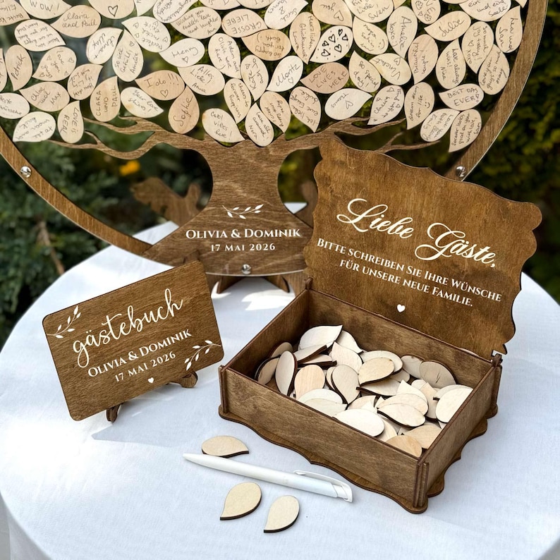 May include: A wooden guest book set for a wedding. The set includes a tree-shaped guest book, a small sign, a box with wooden leaf-shaped tags, and a pen. The tree has leaf-shaped tags with handwritten messages. The box has the text "Liebe G&auml;ste."