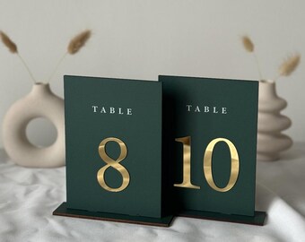 May include: Two dark green table number cards with gold numbers 8 and 10. Each card has the word "TABLE" printed at the top. The cards are on a wooden base, with decorative vases in the background.