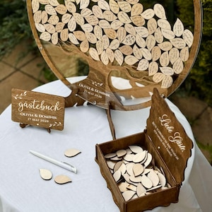 May include: A wooden guest book alternative with a tree design. The tree is filled with wooden leaf-shaped tags for guests to write on. Includes a wooden sign that says "Olivia & Dominik 17 May 2026". A box of blank leaf tags and a pen are also included.