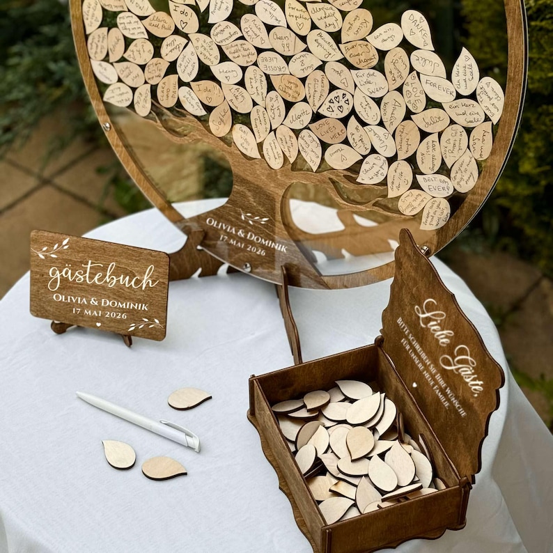 May include: A wooden guest book alternative with a tree design. The tree is filled with wooden leaf-shaped tags for guests to write on. Includes a wooden sign that says "Olivia & Dominik 17 May 2026". A box of blank leaf tags and a pen are also included.