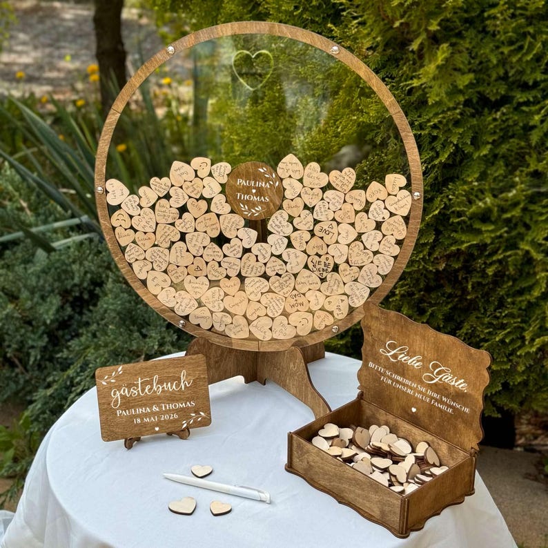 May include: A wooden wedding guest book alternative with a clear circular frame filled with heart-shaped wooden tags. The frame has a wooden base and a small sign that says "Paulina & Thomas". A wooden box and sign are also included.