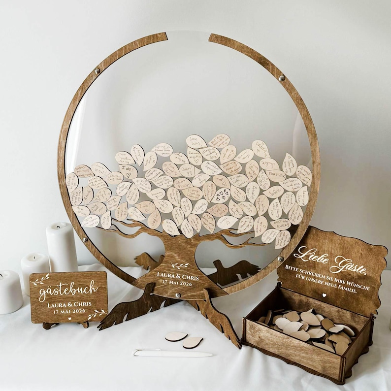 May include: A wooden guest book alternative featuring a tree design with leaf-shaped tags for messages. The tree is set within a circular frame. Includes a wooden box with heart-shaped tags and a sign that reads "Laura & Chris 17 Mai 2026".