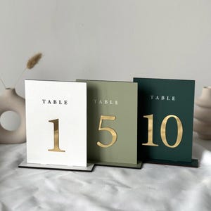 Peut inclure: Trois numéros de table avec des chiffres dorés sur des fonds de couleurs différentes. Les numéros sont 1, 5 et 10. Les fonds sont blanc, vert et vert foncé.