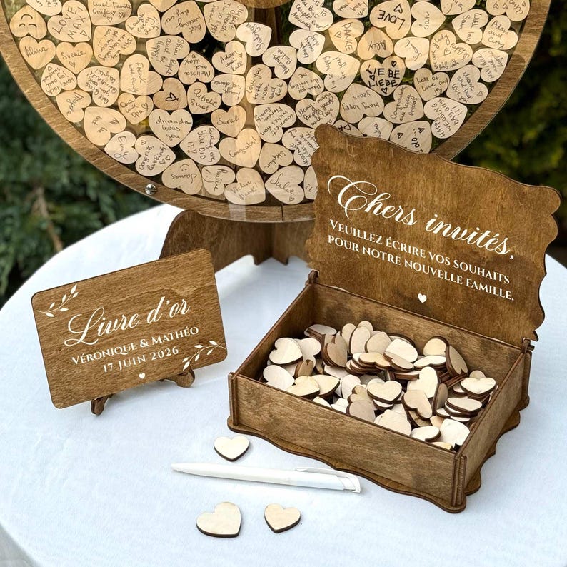 May include: A wooden wedding guest book set. The set includes a large wooden wheel with heart-shaped guest signatures, a wooden box with heart-shaped wooden pieces, and a sign that reads "Livre d'or".