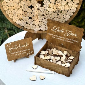 May include: A wooden wedding guest book set. The set includes a large wooden wheel with heart-shaped guest messages, a wooden box filled with heart-shaped wooden pieces, and a sign that reads "g&auml;stebuch" with the date "18 MAI 2026".