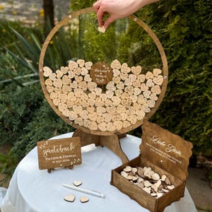 May include: A wooden wedding guest book alternative with a circular frame and heart-shaped wooden pieces. The frame has a clear center and is supported by a wooden stand. A wooden box holds additional hearts. A sign reads "Paulina & Thomas 18 Mai 2026".