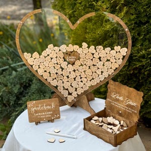 May include: A wooden heart-shaped guest book with a clear front, filled with small wooden hearts. A wooden sign that says "LAURA & CHRIS 17 MAI 2026" is on the left. A wooden box with the words "Liebe Gäste" is on the right.