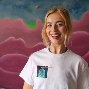 May include: White t-shirt with a graphic of a blue and green design inside a black square, with the word "You niverse" printed next to it. The background features a pink and blue mural.