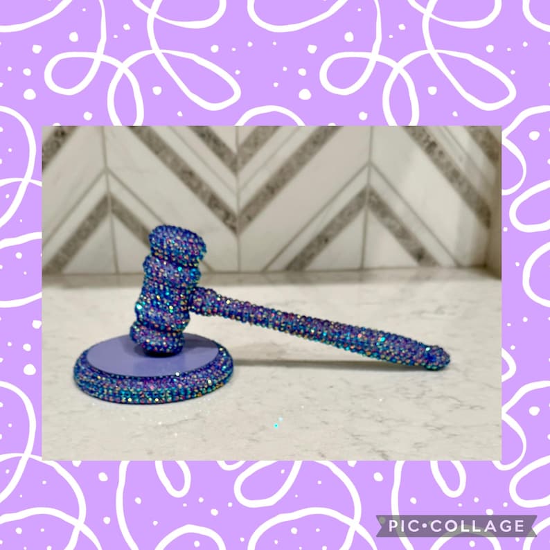 May include: A decorative gavel with a round base. The gavel and base are covered in iridescent rhinestones that reflect blue, purple, and green hues. The base is a solid light purple colour. The gavel is a unique and eye-catching item.