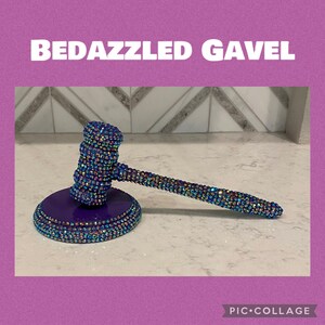 May include: A bedazzled gavel with a purple base and handle, covered in iridescent rhinestones. The text "BEDAZZLED GAVEL" is displayed in white block letters above the gavel.