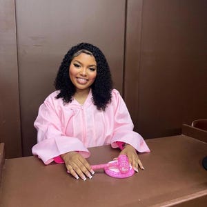 May include: A person wears a pink graduation gown with a zip. A pink gavel and base sit on a brown desk. The person smiles, with curly black hair.