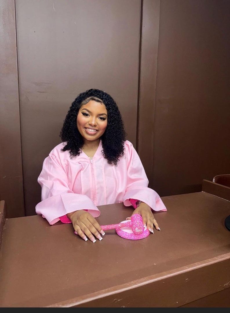 May include: A person wears a pink graduation gown with a zip. A pink gavel and base sit on a brown desk. The person smiles, with curly black hair.