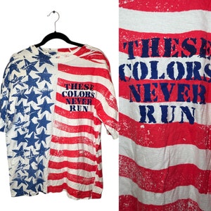 Pode incluir: Uma camiseta branca com um design de bandeira americana vermelha, branca e azul. A camiseta tem o texto "These Colors Never Run" impresso na frente.