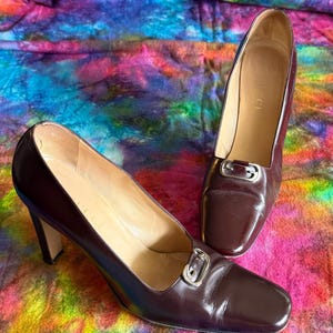 May include: A pair of brown leather high-heeled shoes with square toes and silver-tone buckles. The shoes are displayed on a colorful, tie-dye patterned fabric. The heels are approximately 3 inches high.