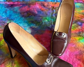 May include: A pair of brown leather high-heeled shoes with square toes and silver-tone buckles. The shoes are displayed on a colorful, tie-dye patterned fabric. The heels are approximately 7.6 cm high.
