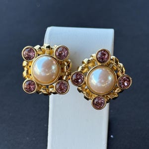 Puede incluir: Un par de pendientes dorados de clip. Cada pendiente presenta una perla falsa grande, redonda y color melocotón, rodeada de cuatro piedras preciosas rosas redondas más pequeñas. Los pendientes tienen forma cuadrada con un diseño dorado retorcido.
