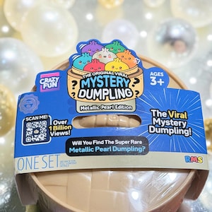 May include: A round, tan plastic container for The Original Viral Mystery Dumpling Metallic Pearl Edition. The blue packaging features colorful cartoon dumplings, a QR code, the text Over 1 Billion Views, and the question Will You Find The Super Rare Metallic Pearl Dumpling? The set is sealed in clear plastic.