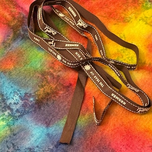 May include: Brown ribbon with white text and stitching. The ribbon reads "HERMES", "THE YEAR OF THE SUN", "ANNEE DU SOLEIL", and "1994". The ribbon is on a colourful, tie-dye background.