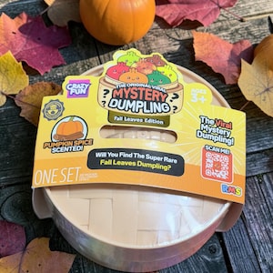 May include: A round, beige plastic container for the Mystery Dumpling Fall Leaves Edition toy. The packaging features a yellow label with the text The Original Viral Mystery Dumpling, Pumpkin Spice Scented, and a QR code for the viral mystery dumpling.
