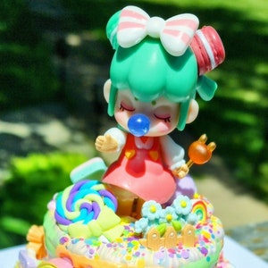 May include: A two-tiered cake decorated with colourful sprinkles, candy, and a small doll with green hair and a pink bow. The cake is topped with a rainbow, a fence, and a ladder.
