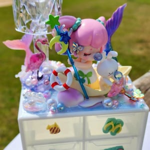 Puede incluir: Un organizador blanco de seis cajones con temática de sirena. El cajón superior presenta una sirena rosa con cola azul, un conejito blanco y un salvavidas. Los otros cajones tienen varias decoraciones coloridas, incluyendo un monstruo verde, un pez amarillo y una tabla de surf verde.