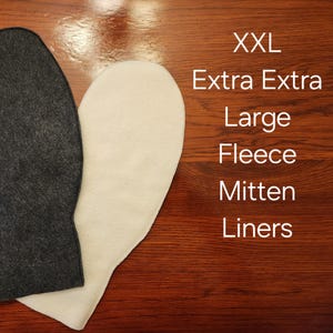 May include: Two XXL fleece mitten liners, one dark gray and one cream-colored, are displayed on a wooden surface. Text on the right side reads "XXL Extra Extra Large Fleece Mitten Liners."
