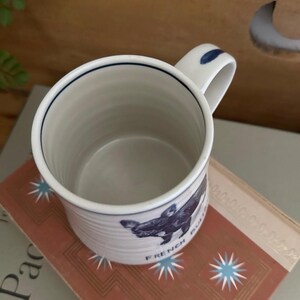 以下が含まれることがあります： 青い縁とハンドルのディテールが施された白いセラミックマグ。マグにはフレンチブルドッグの青いイラストと「FRENCH BULLDOG」の文字が描かれています。マグは花柄のデザインの入った本の上に置かれています。